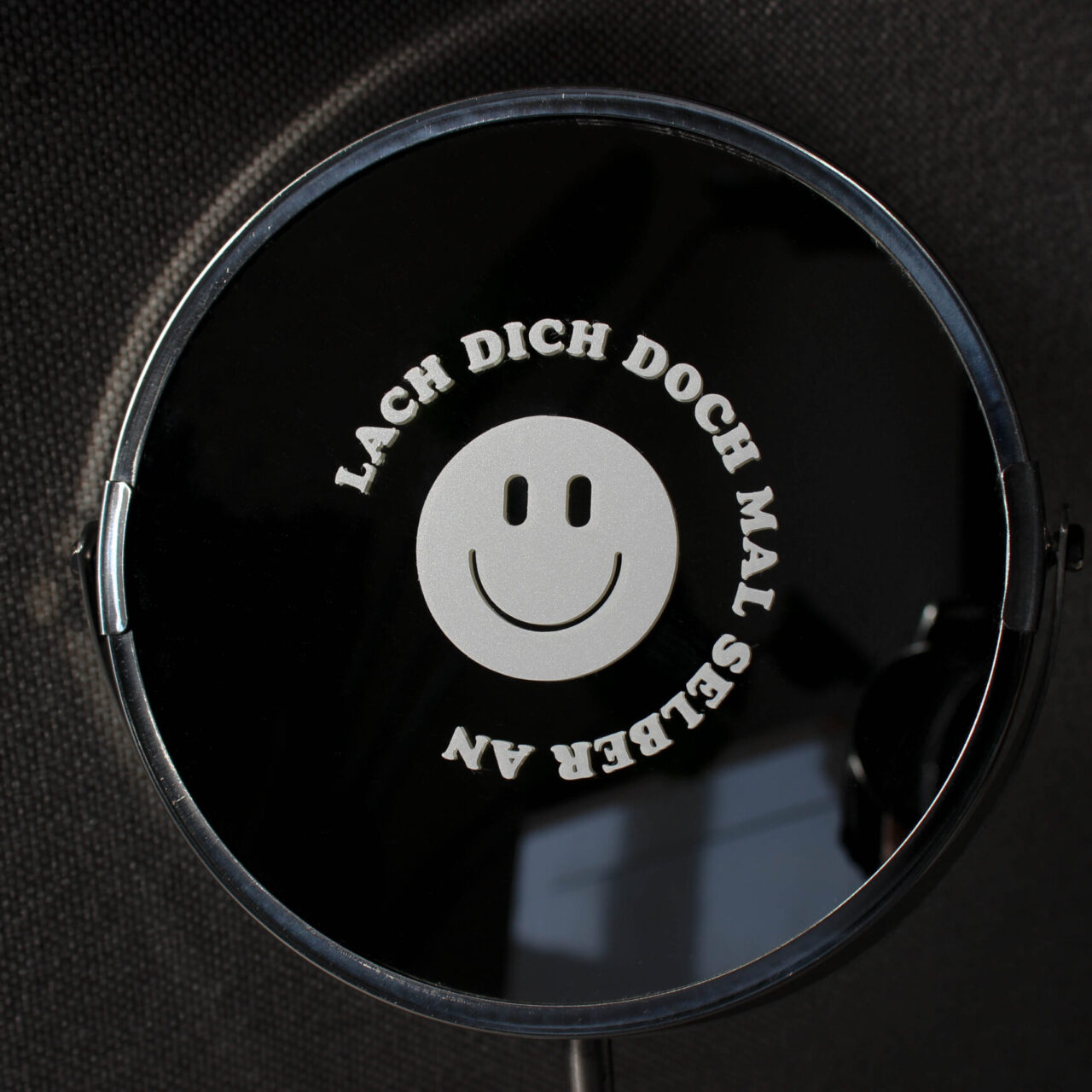 Runder, weißer Sticker "LACH DICH DOCH MAL SELBER AN" mit Smiley auf Spiegel