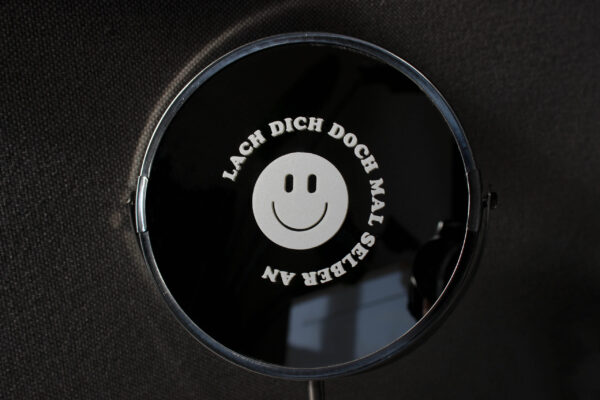 Runder, weißer Sticker "LACH DICH DOCH MAL SELBER AN" mit Smiley auf Spiegel